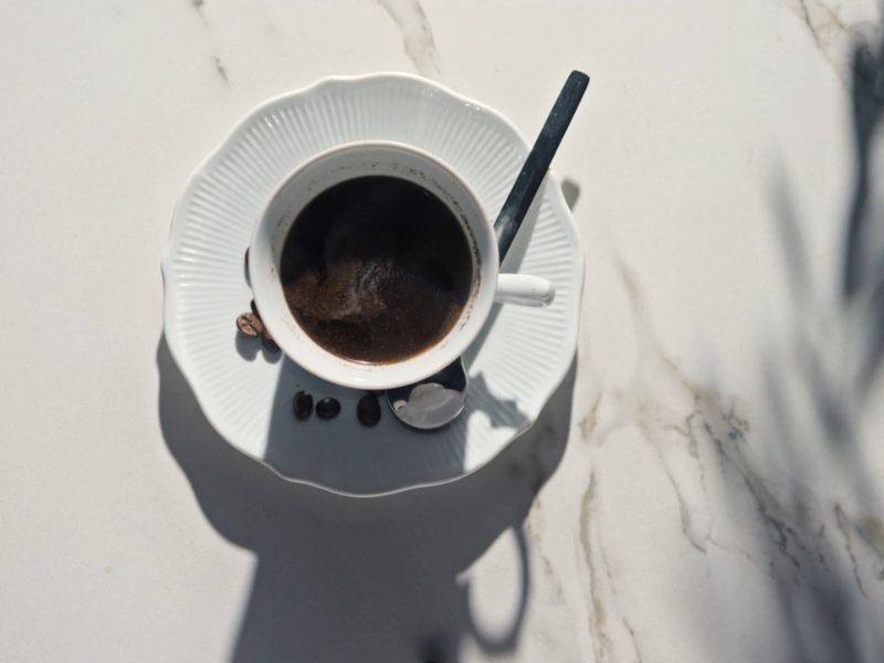 Kafa u beloj šoljici sa tacnom, na belom stolu, par zrna kafe okolo
