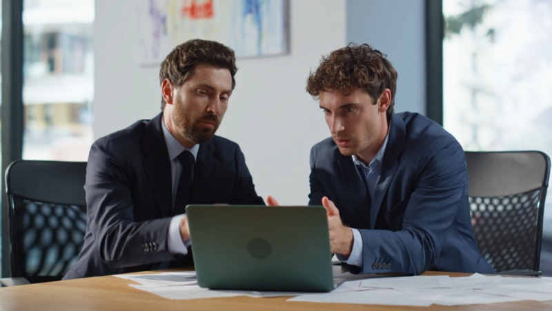 Dva muškarca u odelima sede za stolom i pažljivo analiziraju dokumente na laptopu