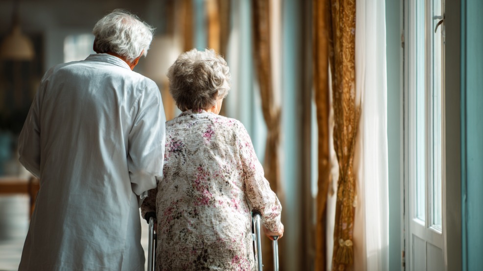 Starija žena uz hodalicu hoda hodnikom, dok je njegovatelj ili zdravstveni radnik prati i pruža podršku