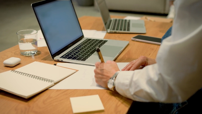 Osoba za radnim stolom zapisuje beleške pored otvorenog laptopa