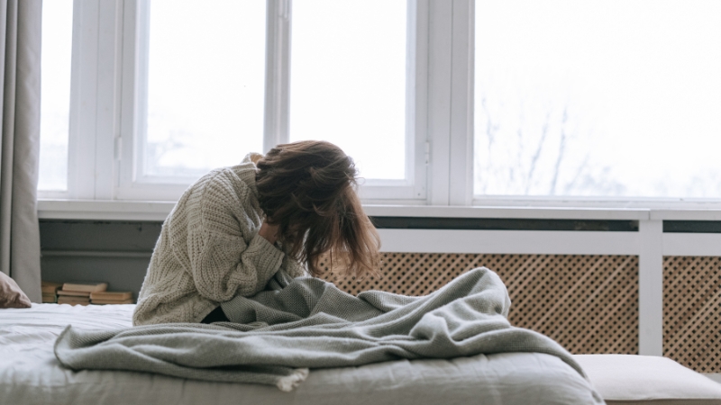 Žena sedi na krevetu i drži glavu rukama, deluje iscrpljeno uprkos snu