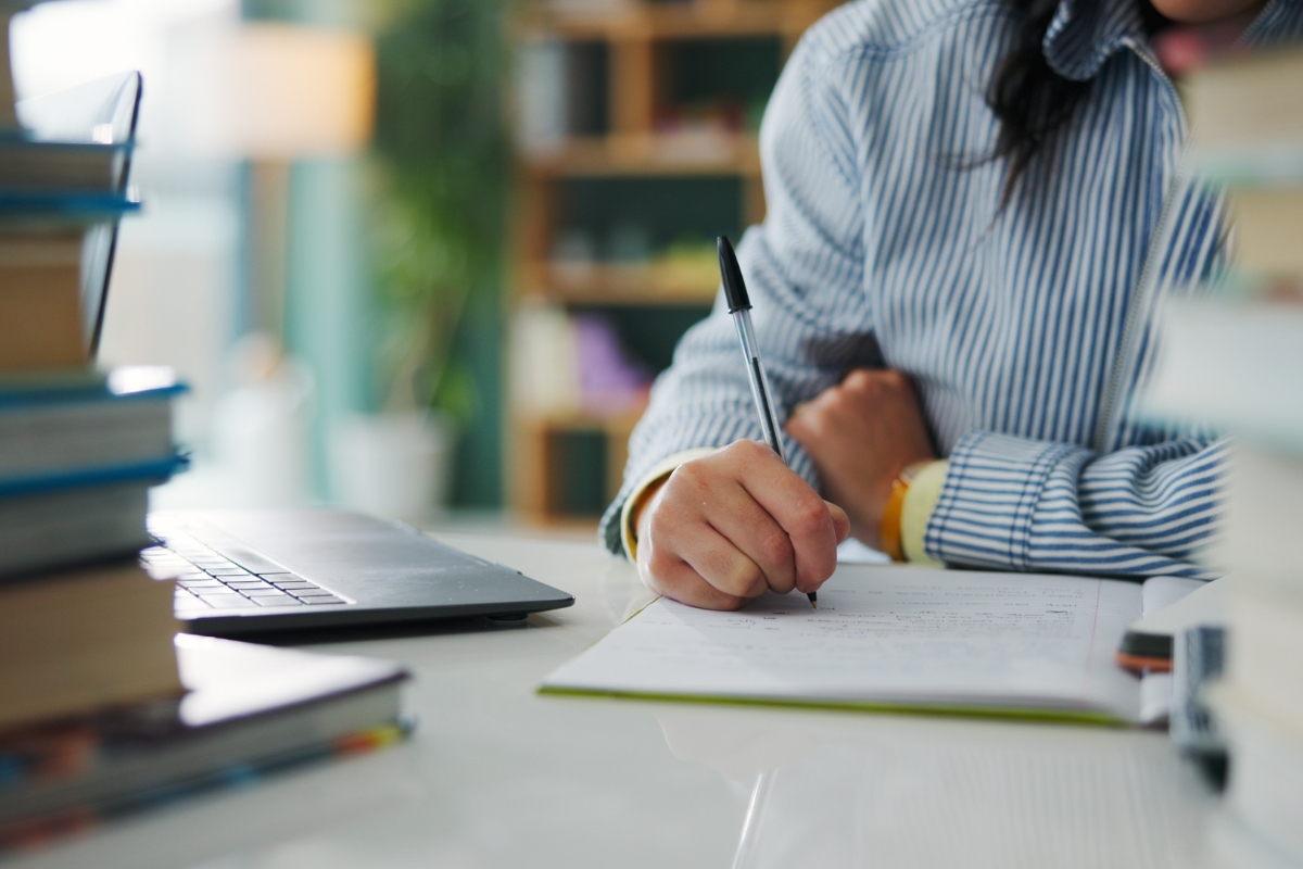 prikaz učenika koji sedi za školskom klupom i piše nešto u knjizi, na stolu knjige i laptop
