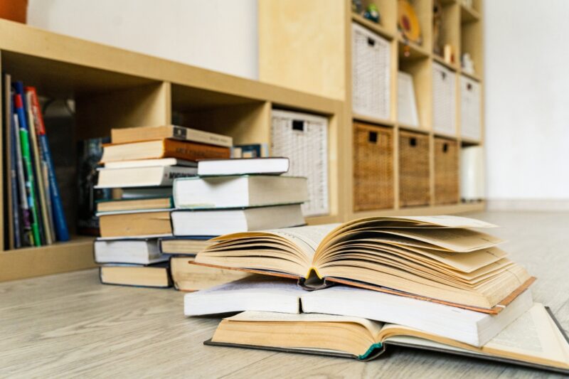 prikaz mnogo knjiga na podu, u pozadini par otvorenih knjiga i polica sa knjigama i fiokama