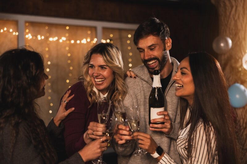 Grupa nasmejanih prijatelja nazdravlja čašama šampanjca u zatvorenom prostoru sa toplim dekorativnim svetlima u pozadini
