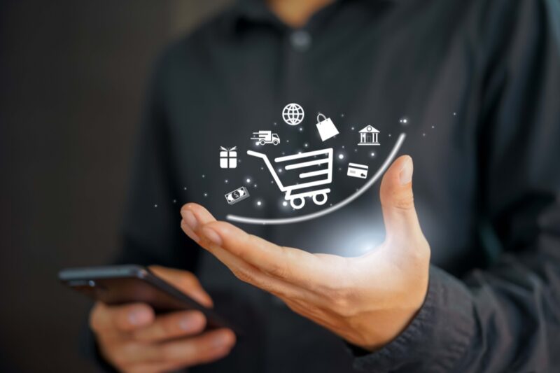 Slika prikazuje osobu koja drži mobilni telefon dok iznad šake lebdi digitalni simbol kolica za kupovinu, okružen ikonama povezanim sa online prodajom, kao što su paketić, kamion za dostavu, novčanica, kartica i simbol interneta, što ilustruje koncept e-trgovine i digitalne kupovine.