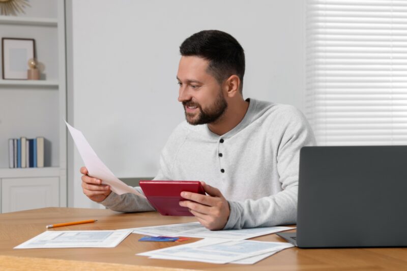 momak gleda u tablet i papire na stolu, pored njega laptop