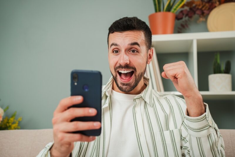 Muškarac zadovoljno gleda rezultat cashback promocije na mobilnom telefonu nakon sportske opklade ili kazino igre, povezan sa temom obrtnih uslova i isplate bonusa