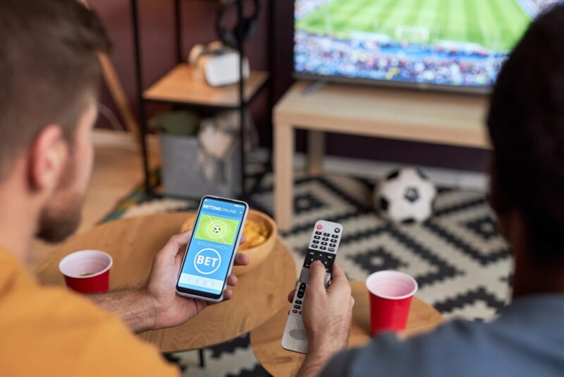 Dve osobe gledaju fudbalsku utakmicu dok jedna koristi mobilnu aplikaciju za sportsko klađenje, povezano sa izborom cashback ponuda i odgovornim igranjem