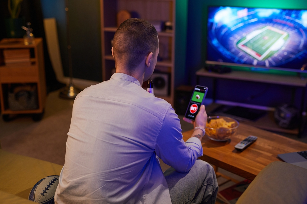 Muškarac sedi na kauču, gleda fudbalsku utakmicu na televizoru i drži telefon u ruci