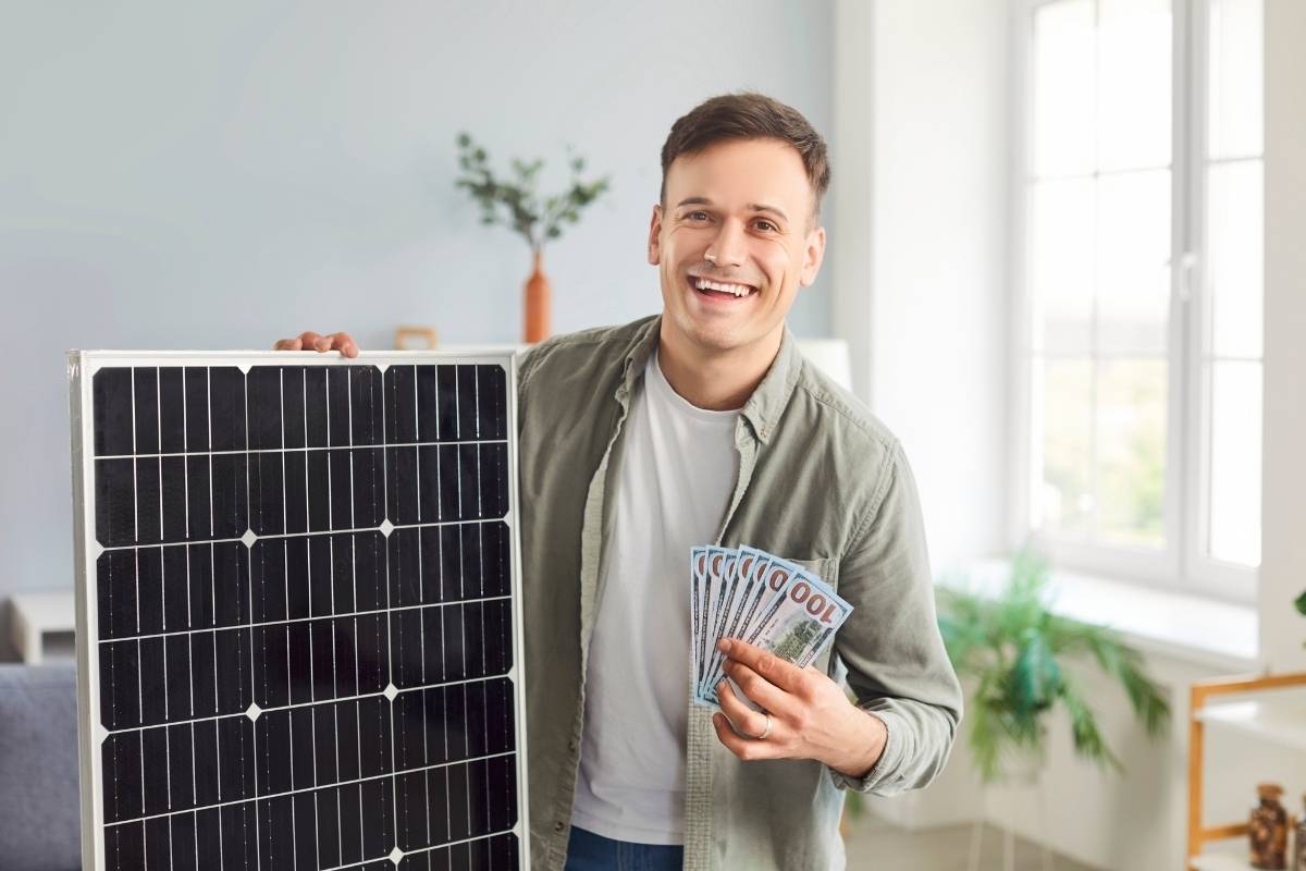 osoba u jednoj ruci drži solarni panel a u drugoj novac i smeje se